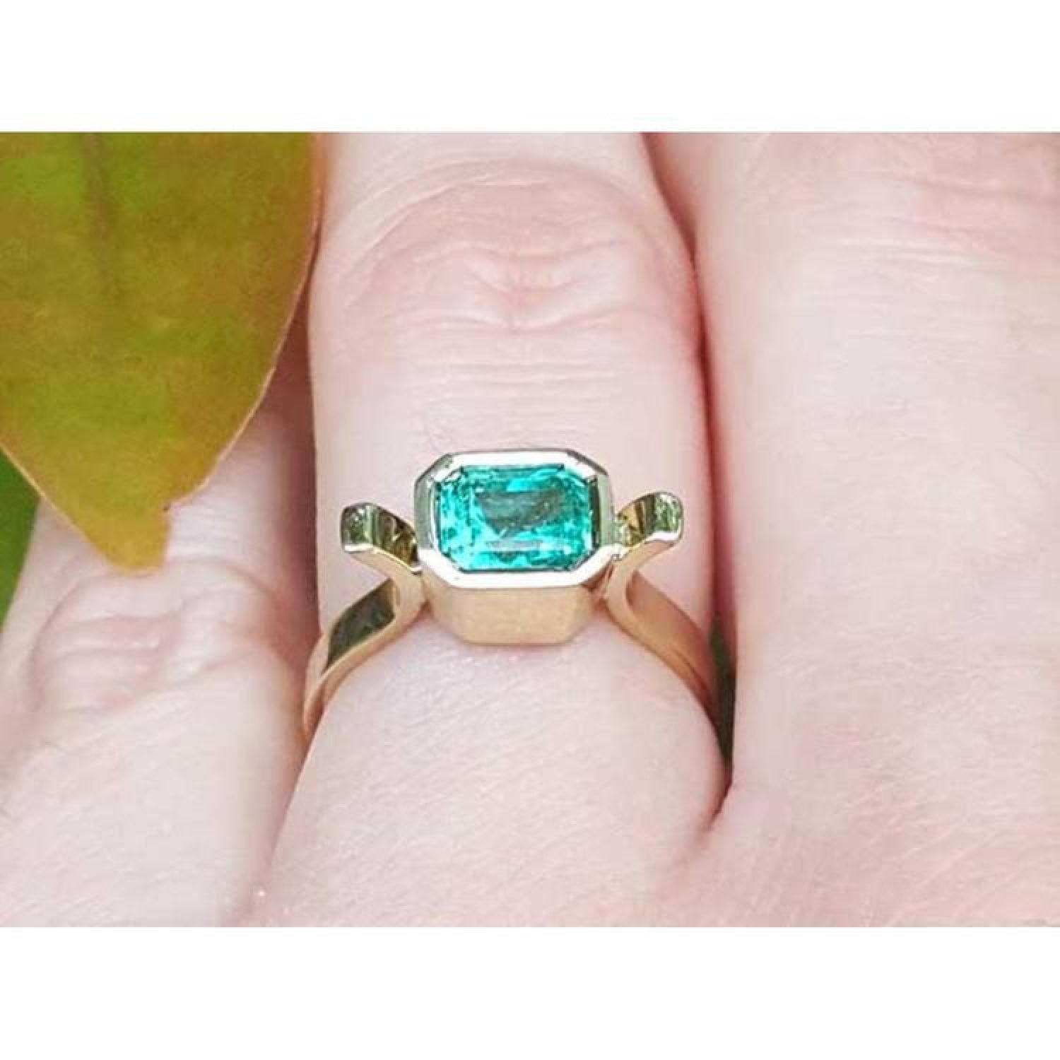 Bezel-set emerald cut Colombian emerald ring in 14K yellow gold on a finger.
