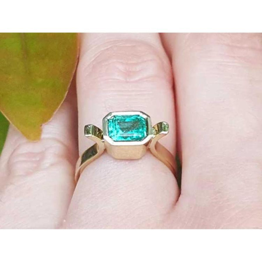 Bezel-set emerald cut Colombian emerald ring in 14K yellow gold on a finger.