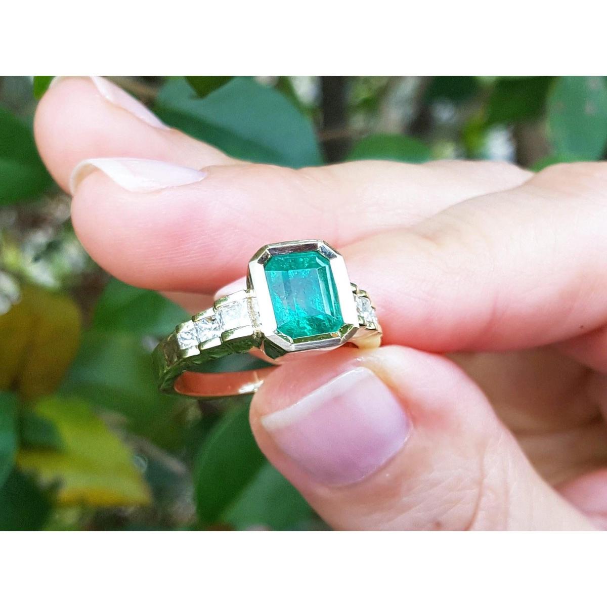 A natural Colombian emerald ring for women held between fingers with a blurred green leafy background.