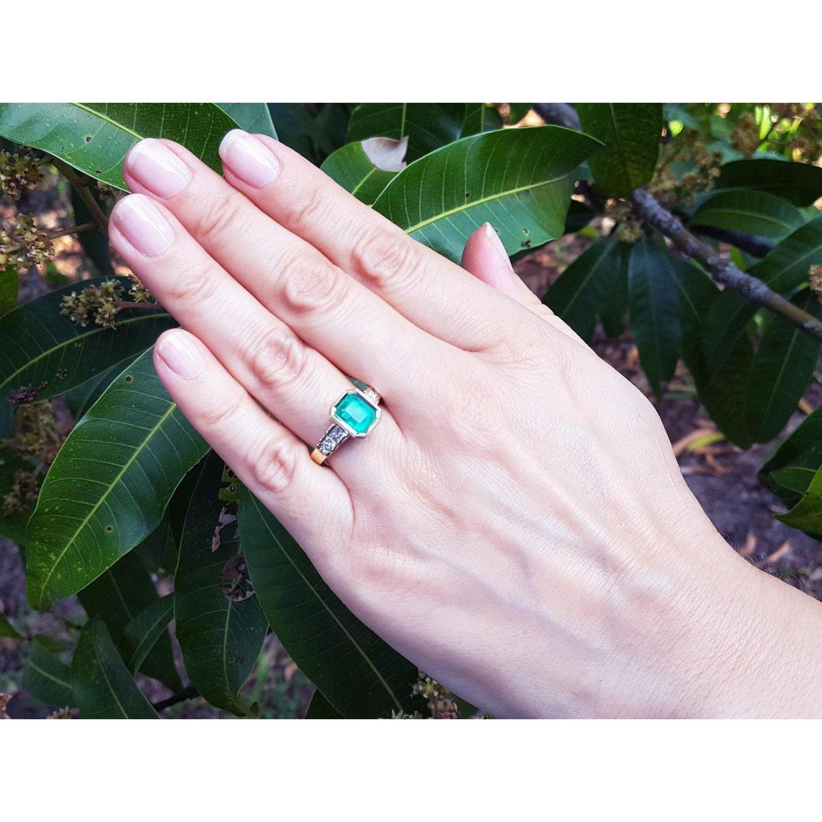 Hand wearing a bezel set emerald ring with accent princess cut diamonds against a leafy background.