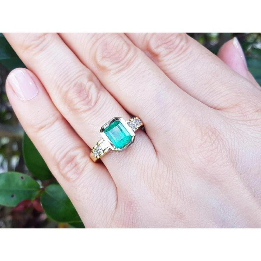 18K gold ring with a 2.10 ct rectangular emerald and bezel-set princess cut diamonds on a hand.