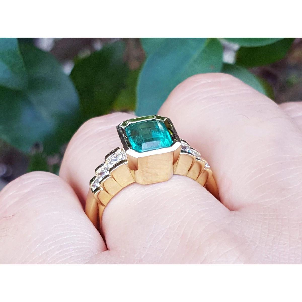 A women's Colombian emerald ring bezel set on a finger against a blurred green leafy background.