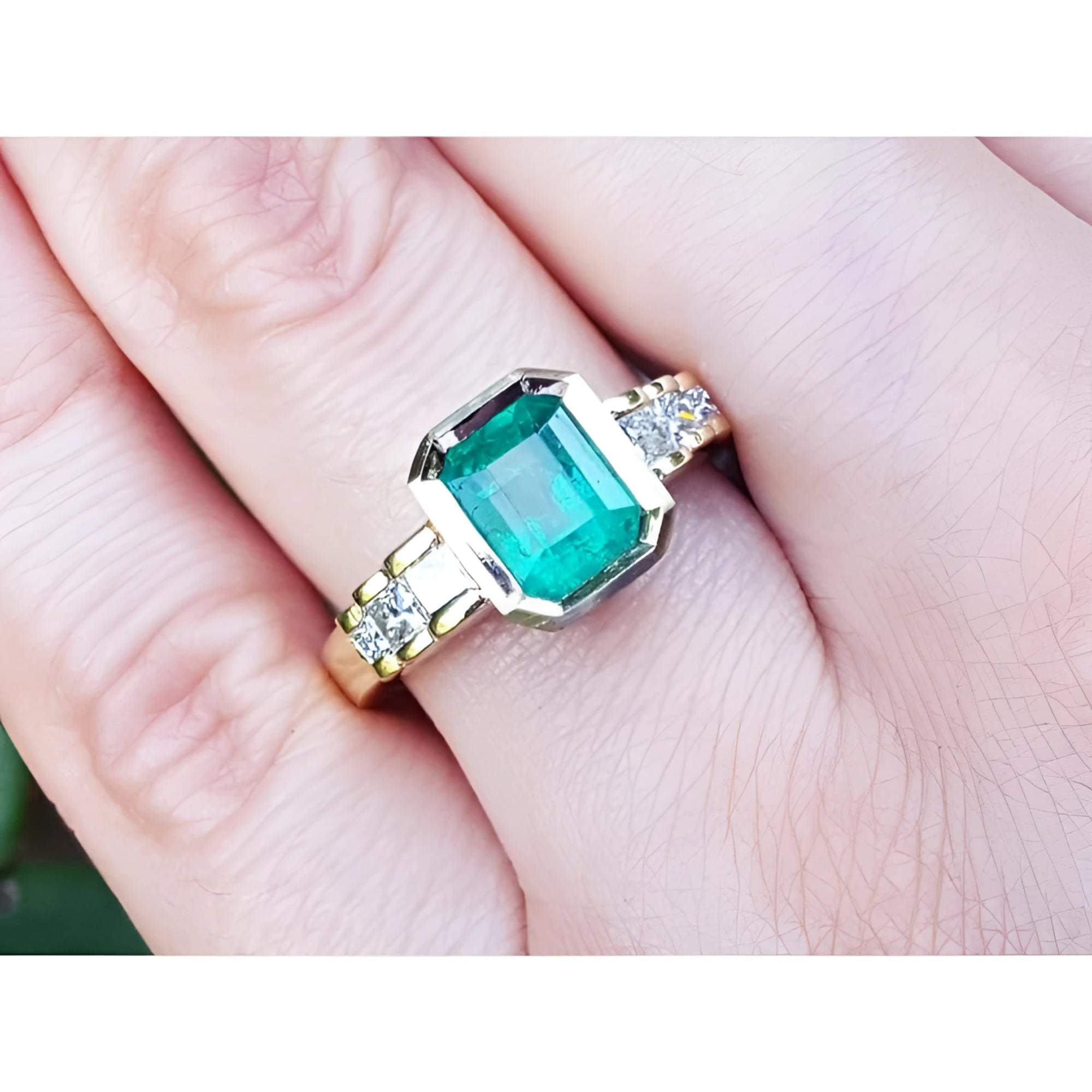 A bezel set emerald ring with accent side princess cut diamonds crafted in 18k yellow gold on a finger.