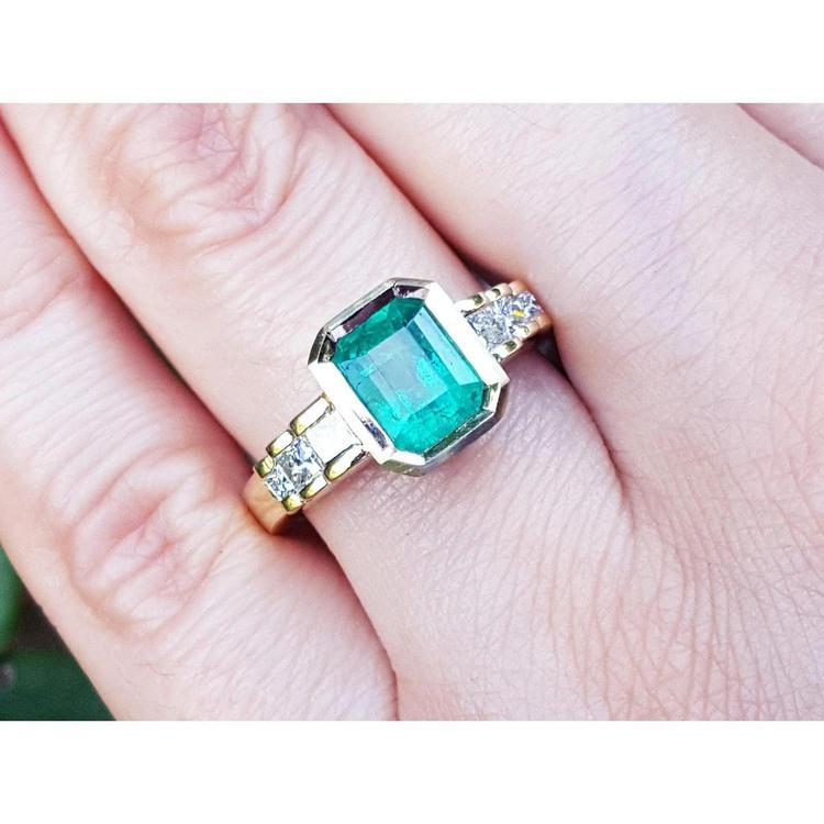 A bezel set emerald ring with accent side princess cut diamonds crafted in 18k yellow gold on a finger.