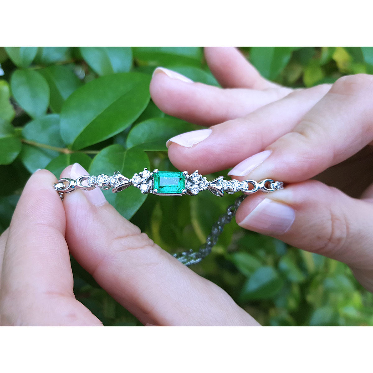 A white gold bracelet with an emerald gemstone held by hands against a green leafy background.