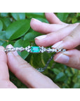 A white gold bracelet with an emerald gemstone held by hands against a green leafy background.
