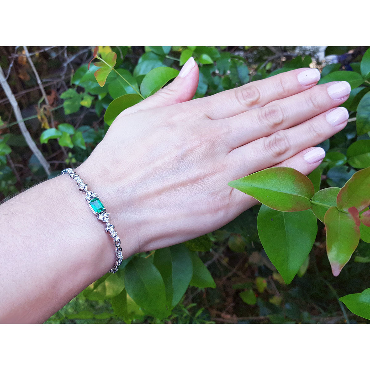 Hand wearing a bracelet with emerald and diamonds against a green leafy background.