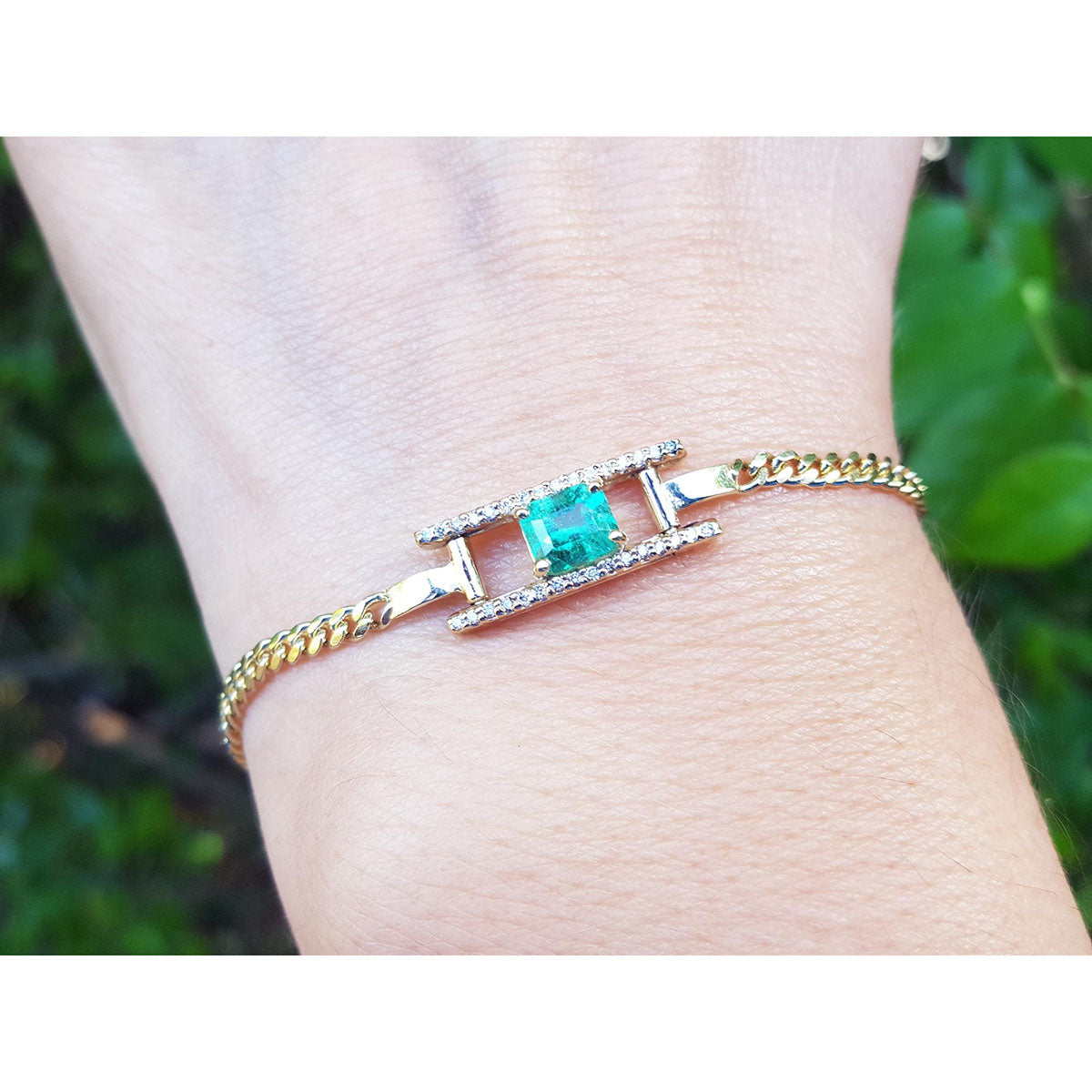 Gold bracelet with a square natural emerald on a wrist against a blurred green background.