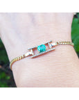 Gold bracelet with a square natural emerald on a wrist against a blurred green background.