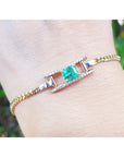 Gold bracelet with a square natural emerald on a wrist against a blurred natural background.