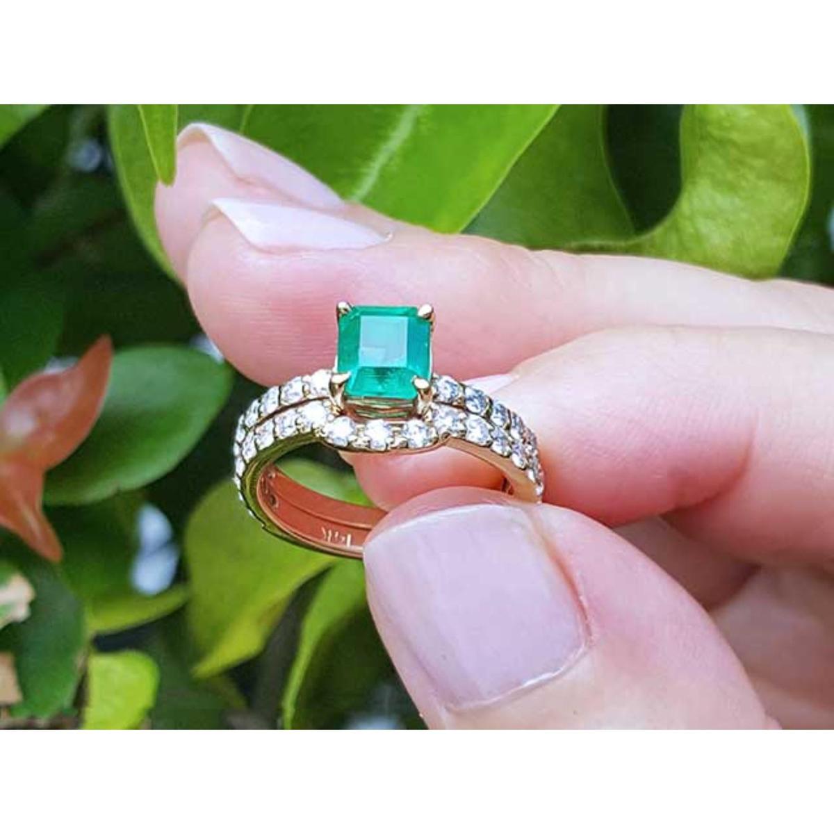 An emerald ring with matching band set held between fingers against a green leafy background.