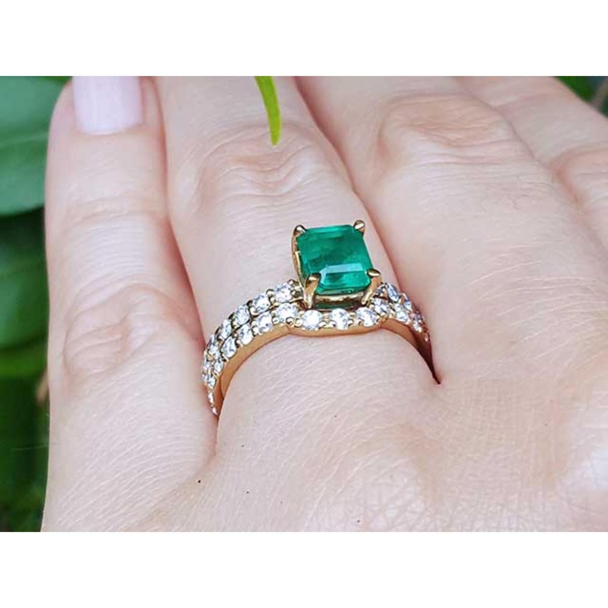 A yellow gold emerald bridal ring with the matching band set on a finger against a blurred natural background.