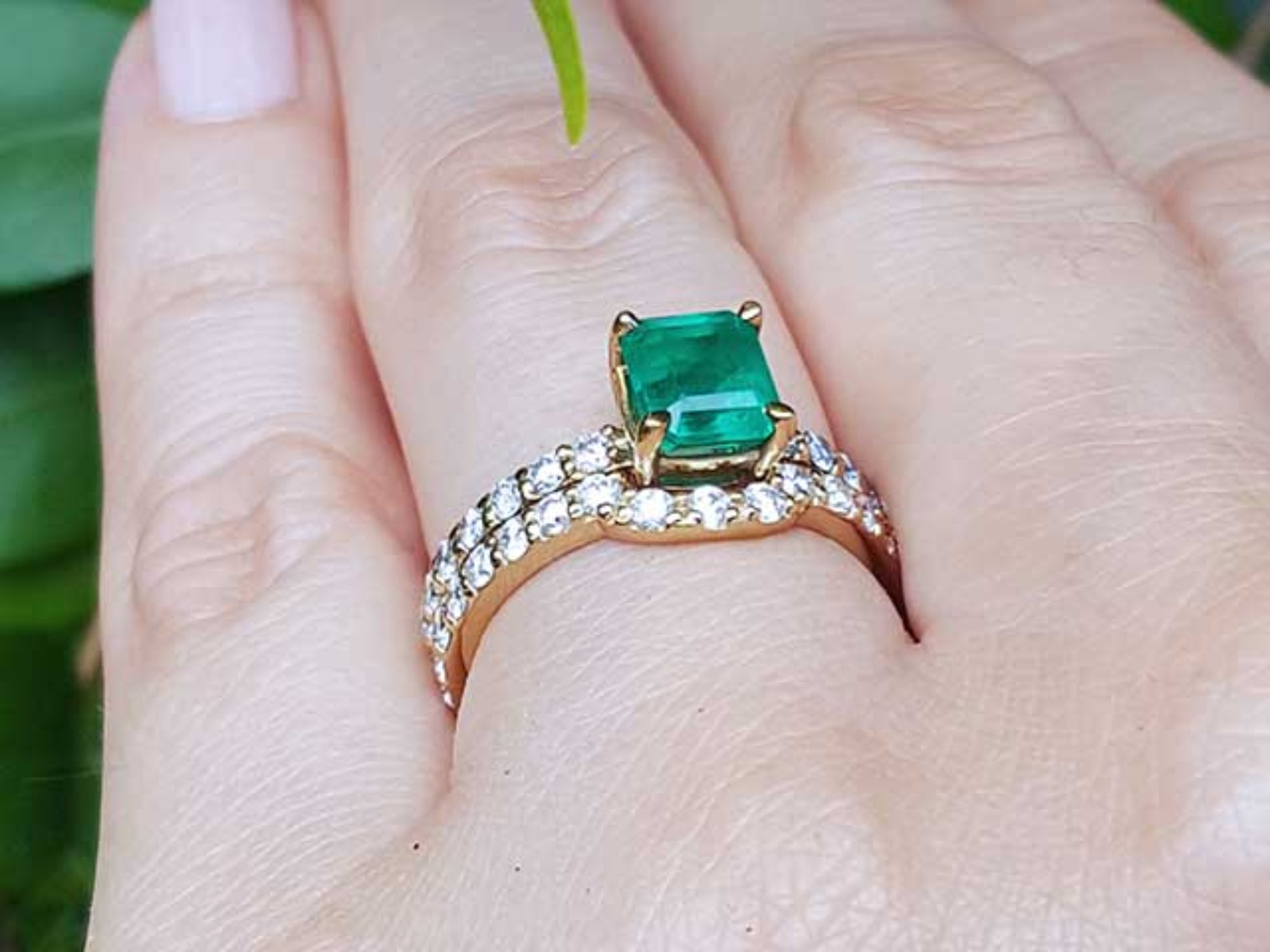 A yellow gold emerald bridal ring with the matching band set on a finger against a blurred natural background.