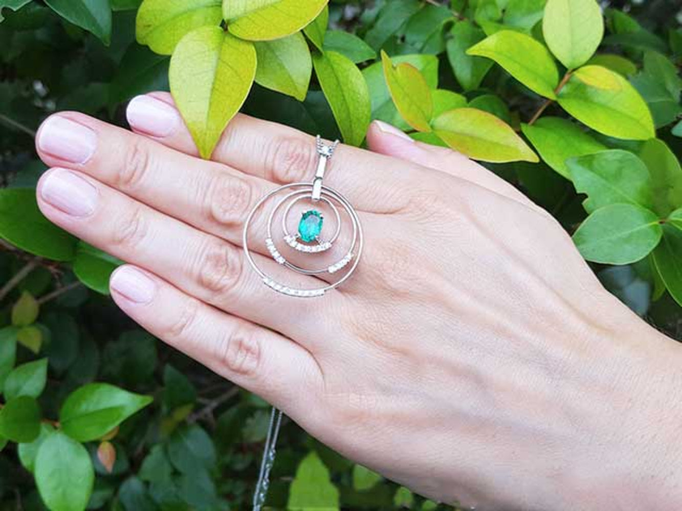 Hand displaying a circle of life pendant with an oval cut emerald against a green leafy background.