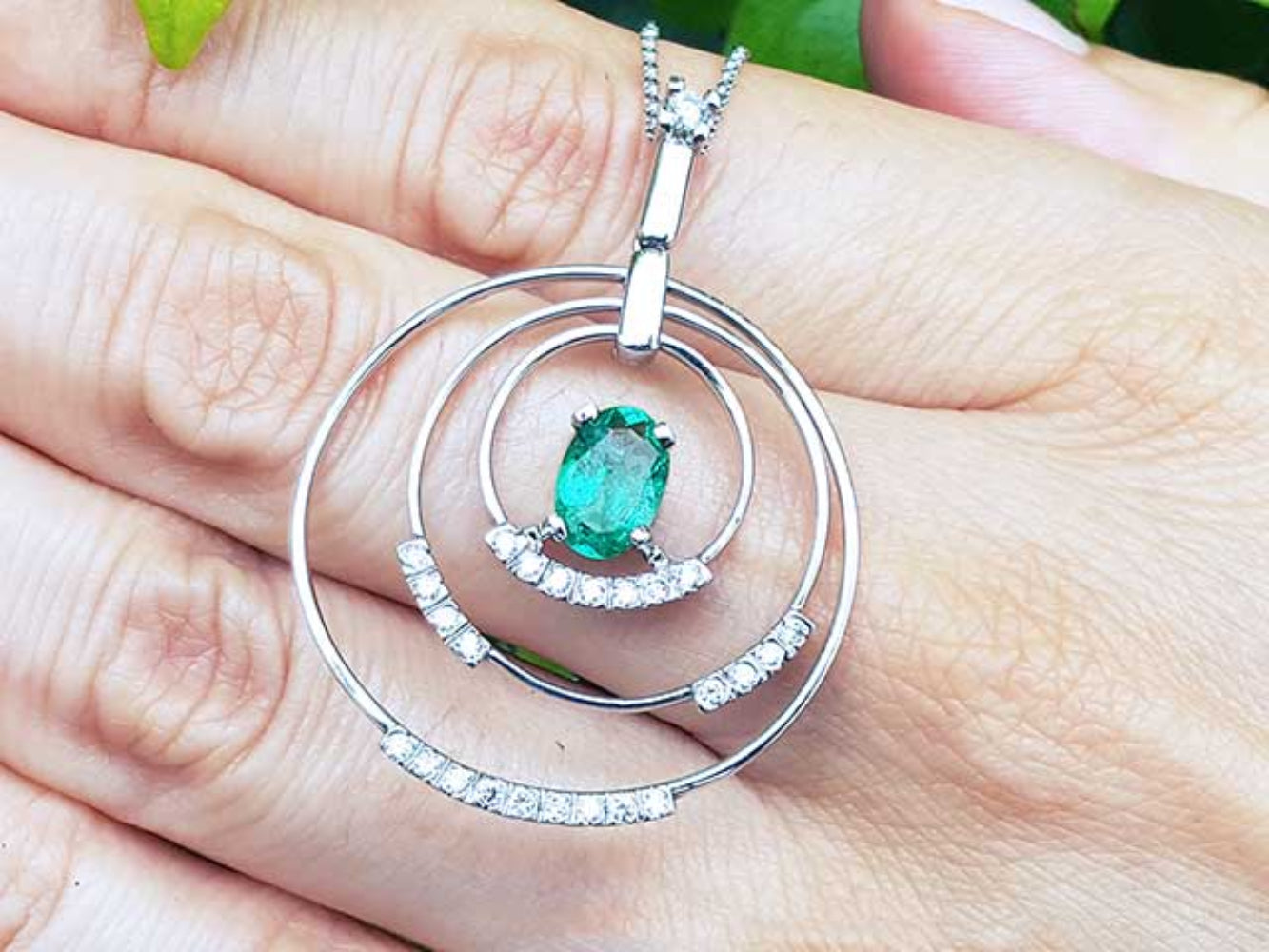 A white gold circle of life emerald pendant with diamonds held on a hand.