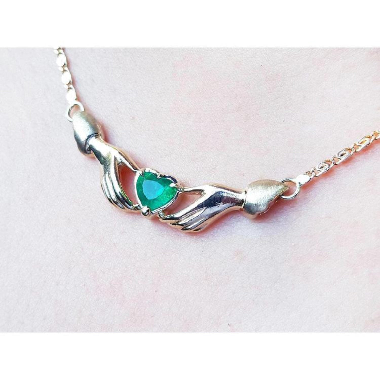 14K yellow gold Claddagh necklace with a heart-cut emerald held by two hands, worn on a neck.