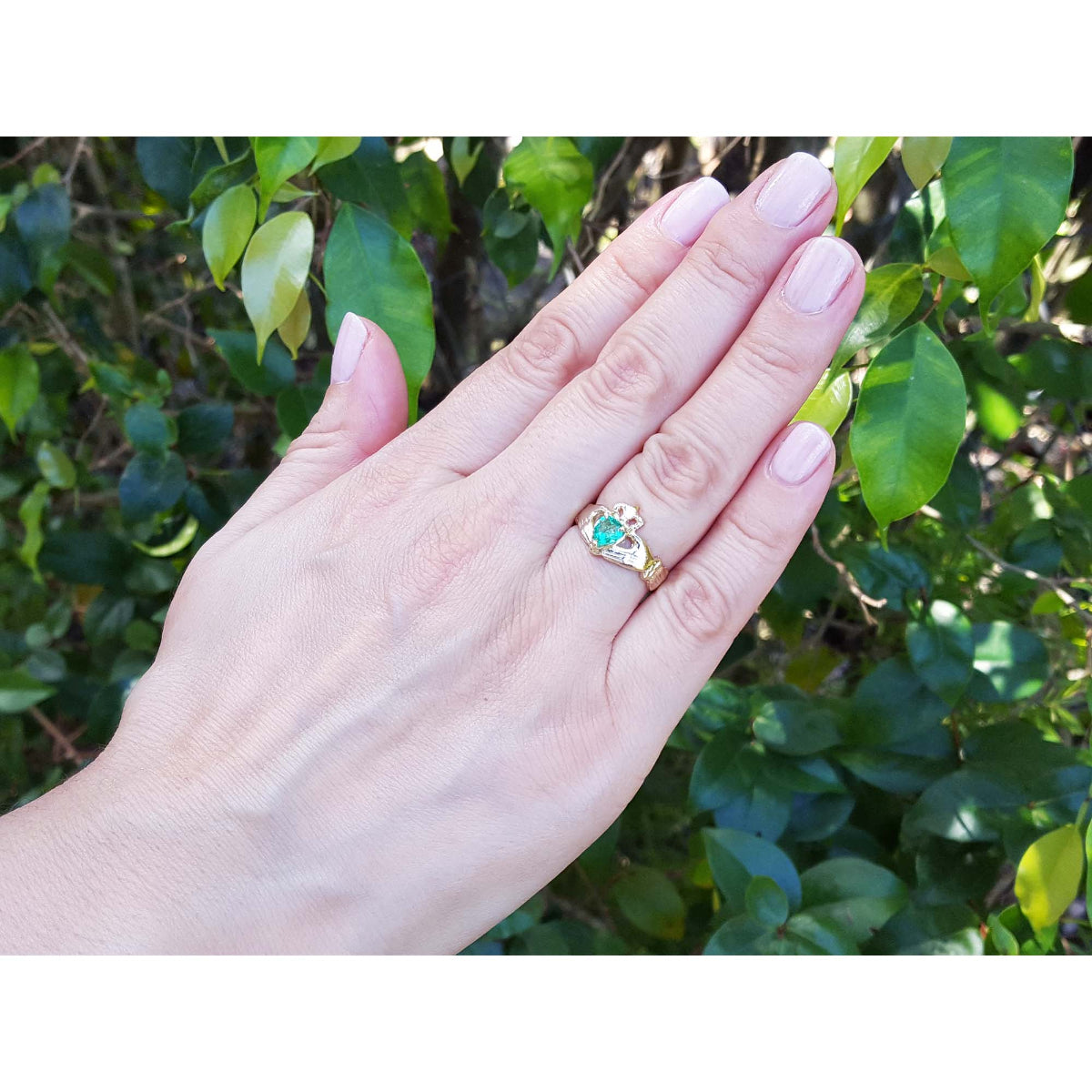 Hand wearing a 14k yellow gold emerald claddagh ring against a leafy background.