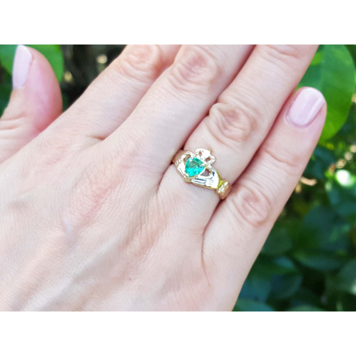 Hand wearing an emerald claddagh ring crafted in 14k yellow gold against a blurred green background.