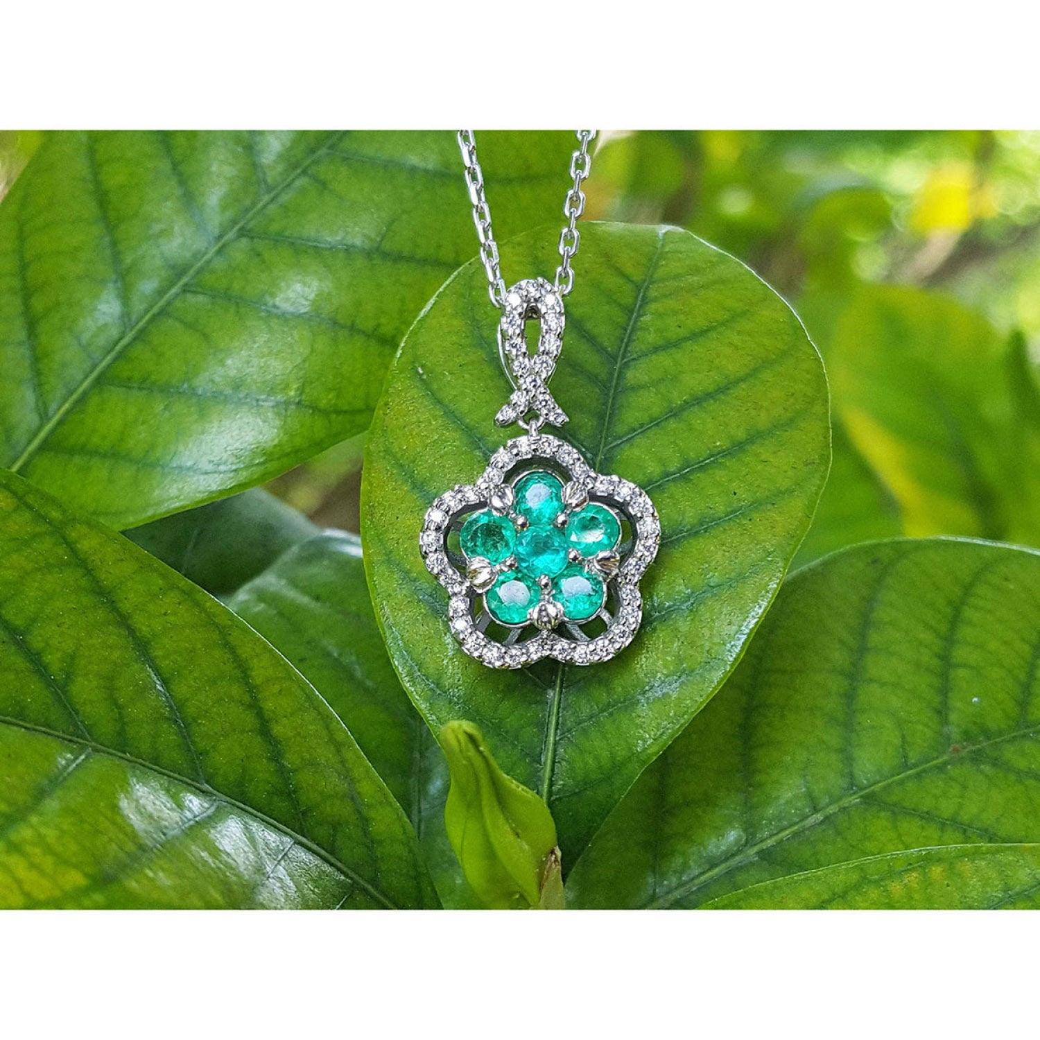 14K white gold emerald cluster necklace with five petals, surrounded by diamonds, on a green leaf.