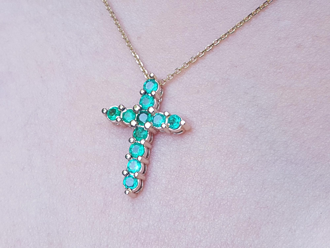 Cross-shaped necklace with natural emeralds on a light pink skin background.