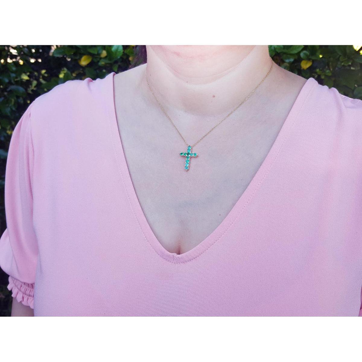 A woman wearing a pink top with a necklace featuring an emerald cross pendant.