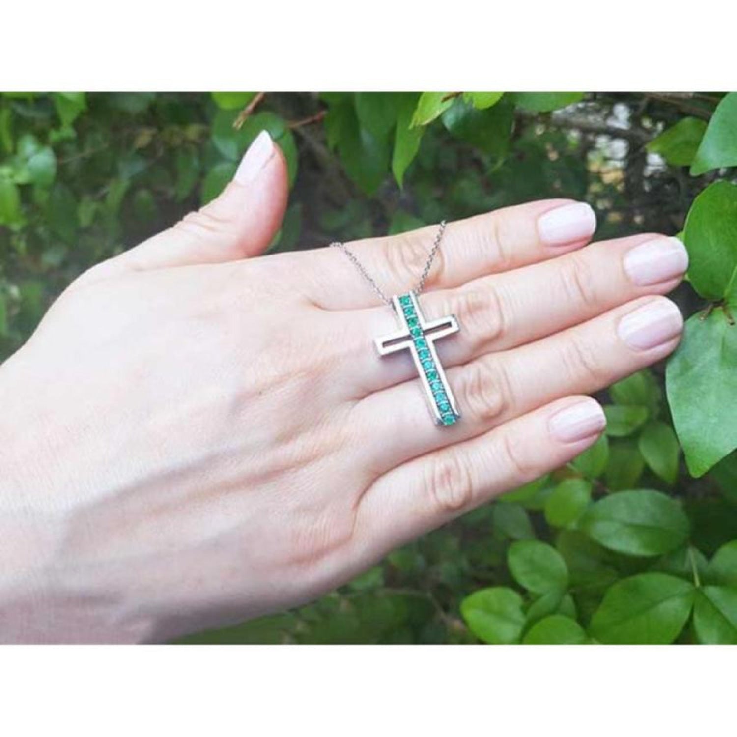 14K white gold cross pendant with prong-set round Colombian emeralds on a delicate chain, held in a hand.