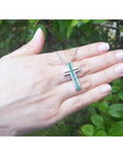 14K white gold cross pendant with prong-set round Colombian emeralds on a delicate chain, held in a hand.