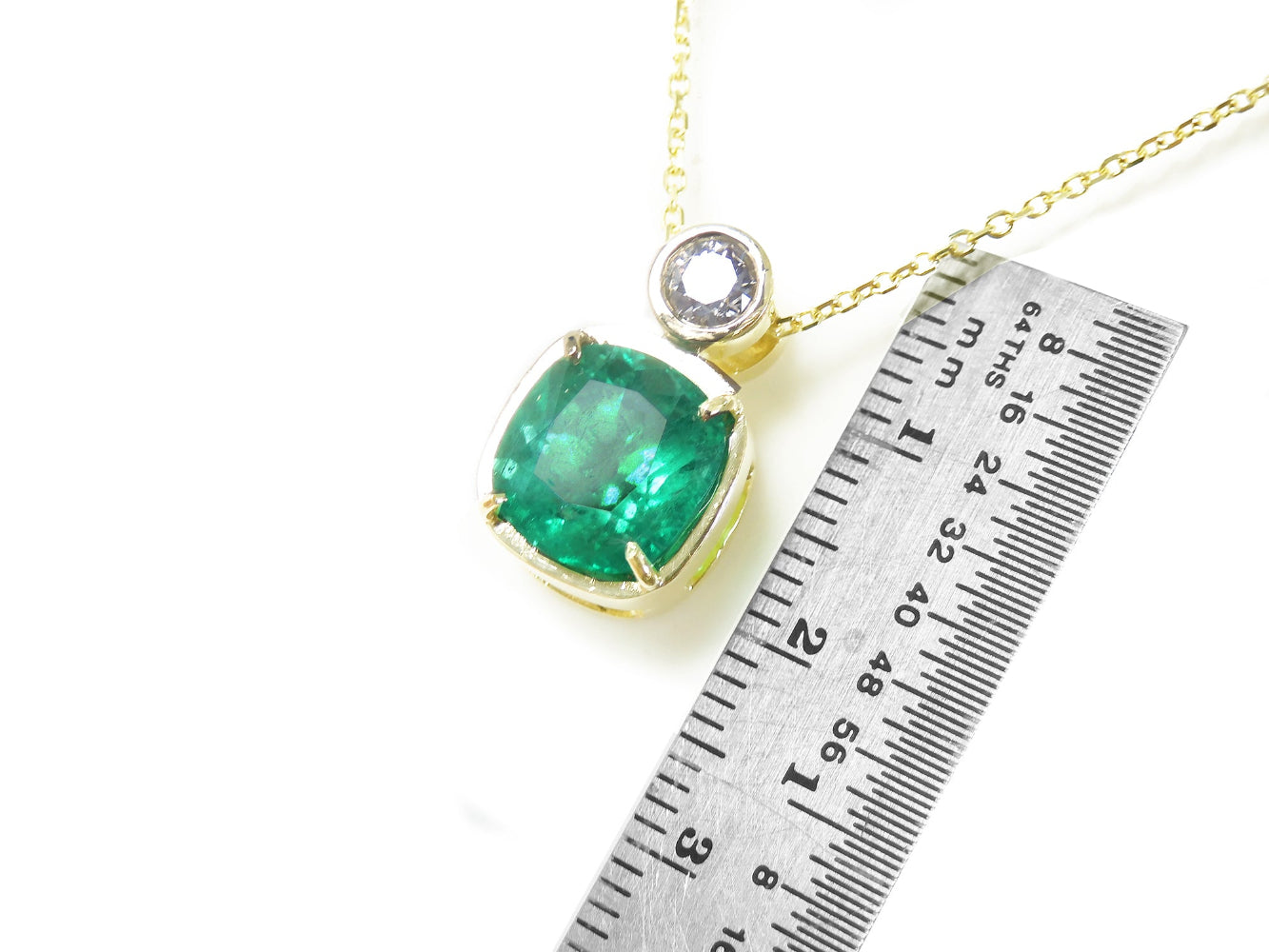 A yellow gold cushion cut emerald and diamond pendant next to a ruler for scale on a white background