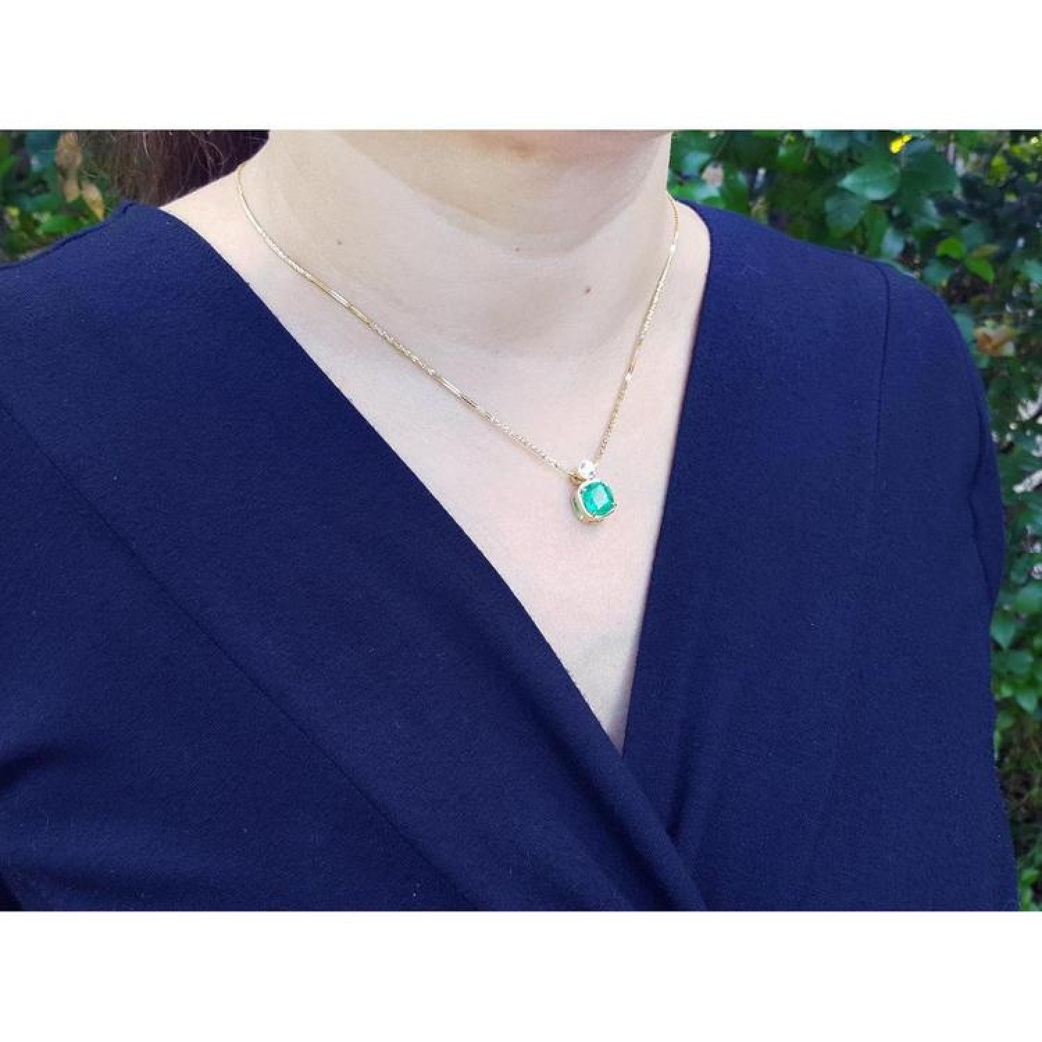 Woman wearing a vibrant deep bluish-green emerald cushion cut pendant in 18K yellow gold.