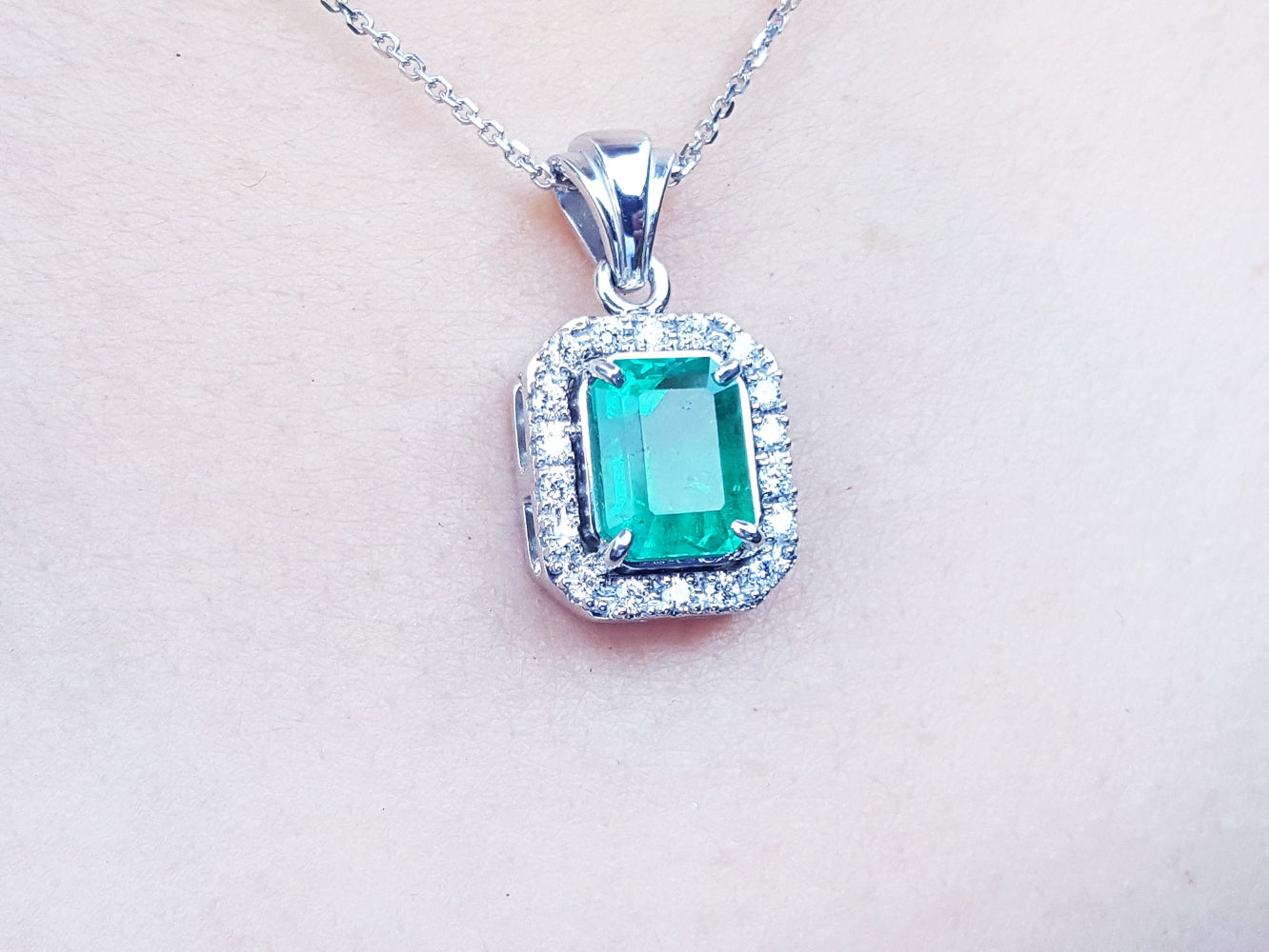 A white gold emerald pendant with a halo diamond on a light pink background.