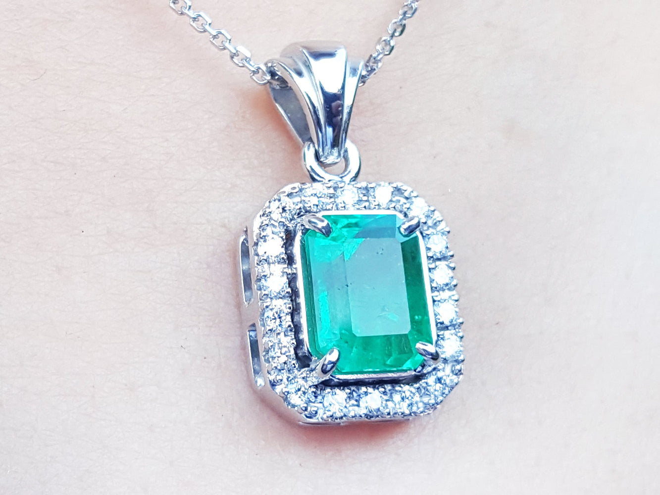 A close-up of an emerald gemstone pendant with white gold and diamonds frame on a light pink background.