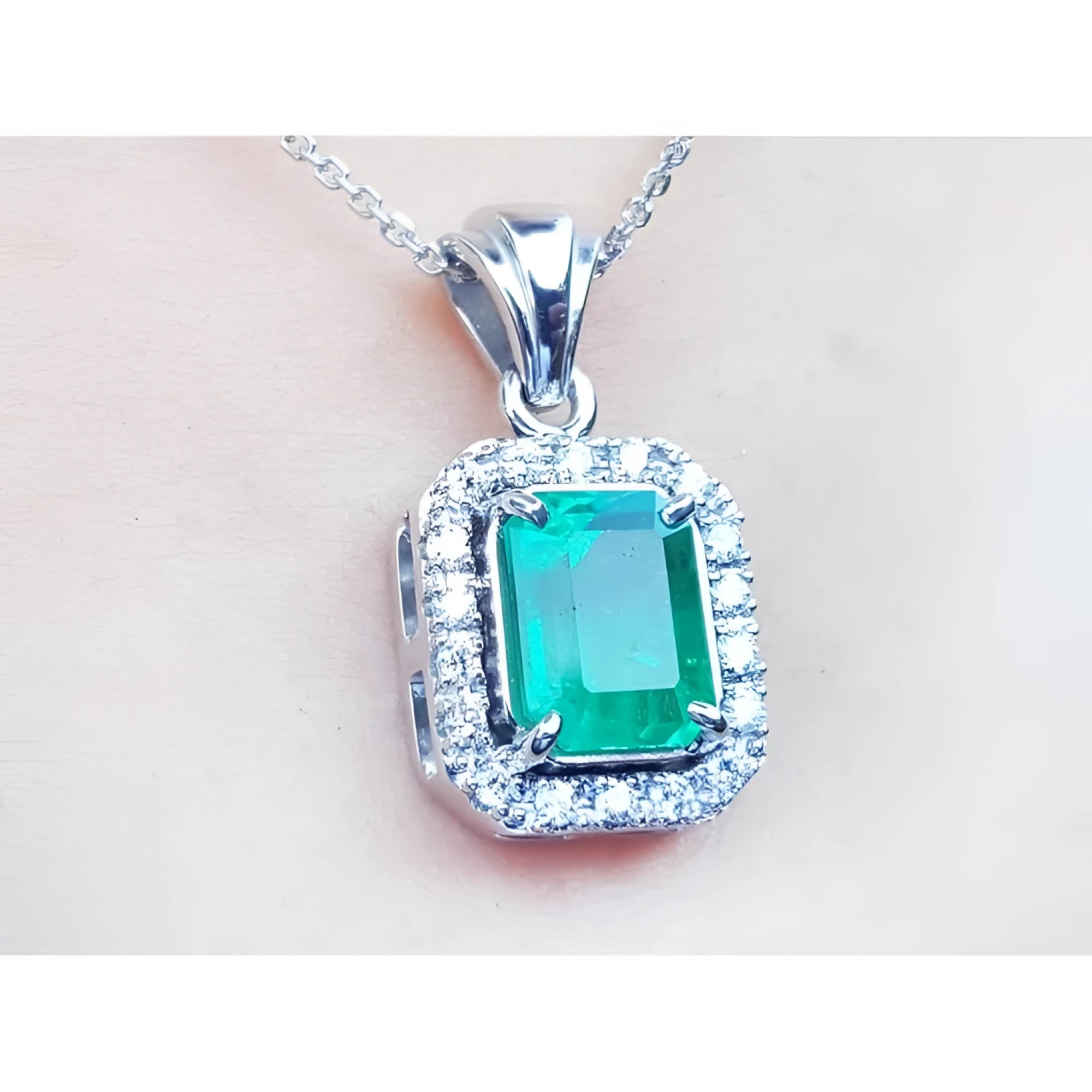 A close-up of an emerald gemstone pendant with white gold and diamonds frame on a light pink background.