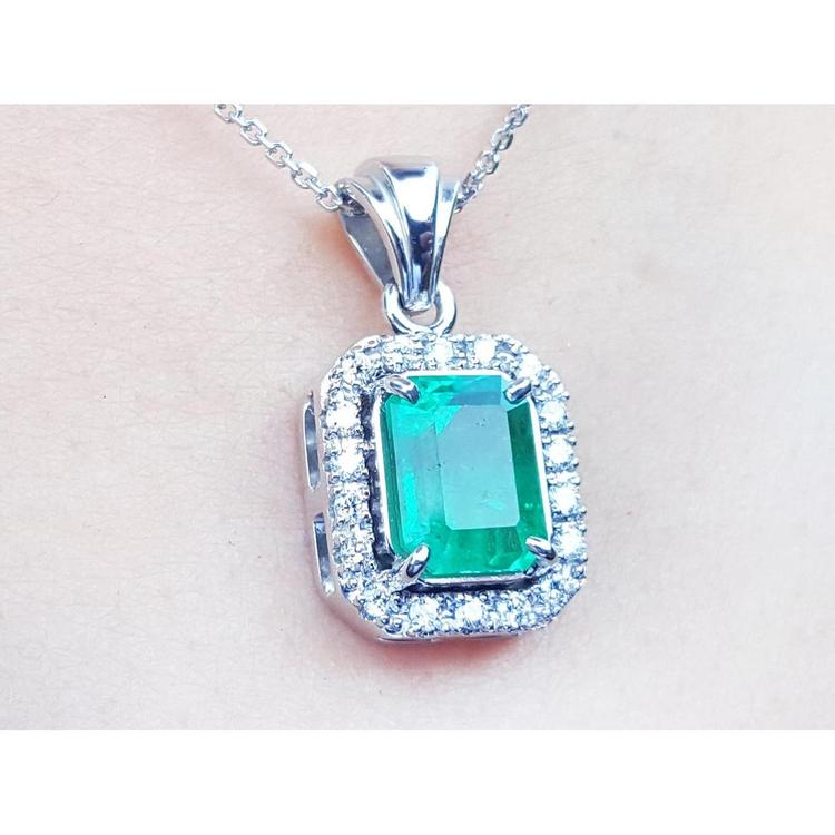 A close-up of an emerald gemstone pendant with white gold and diamonds frame on a light pink background.