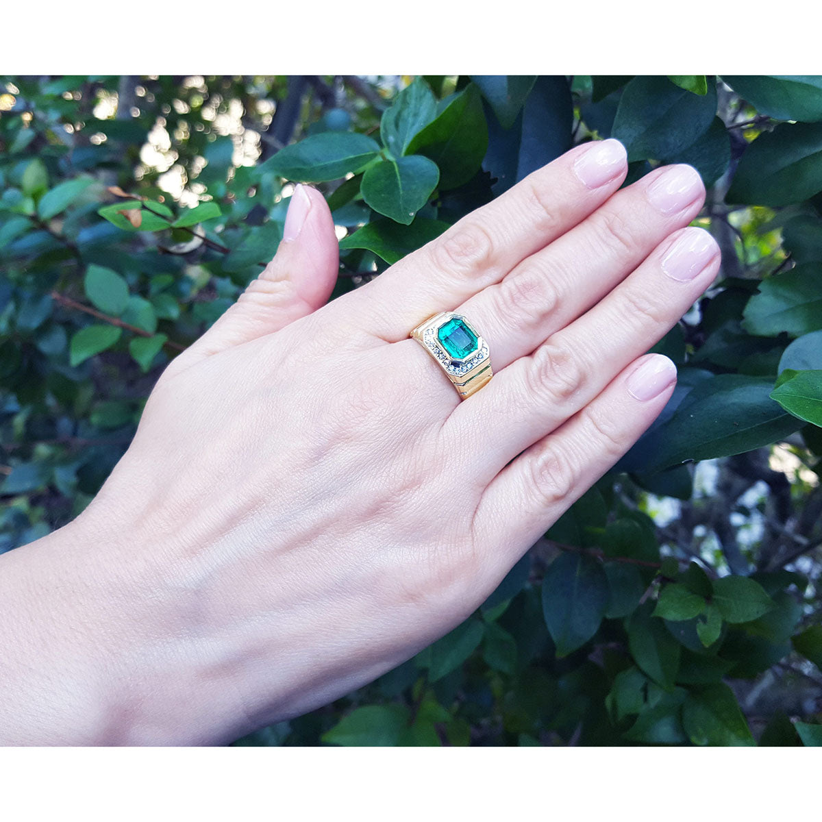 Hand wearing a mens gold ring with a rectangular emerald against a leafy background.
