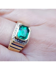 An 18k yellow gold men’s emerald ring with sparkling accent diamonds resting on a finger.