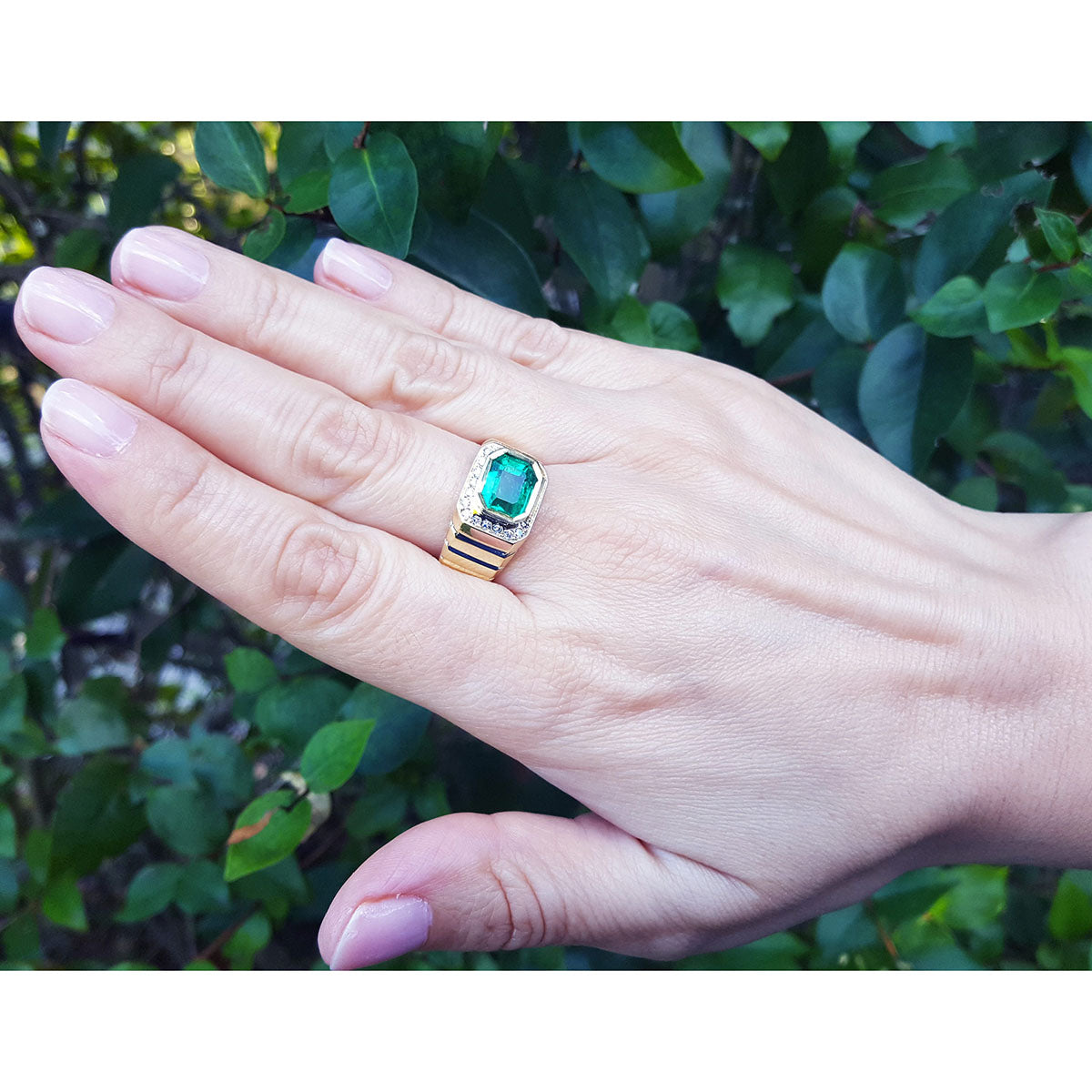 Hand wearing a mens ring with a natural colombian emerald against a green leafy background.