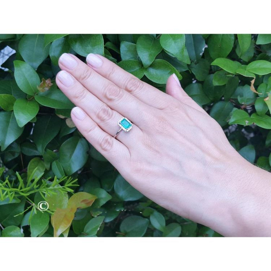 Emerald-cut Muzo emerald halo diamond ring in 14K white gold on a hand with green foliage background.