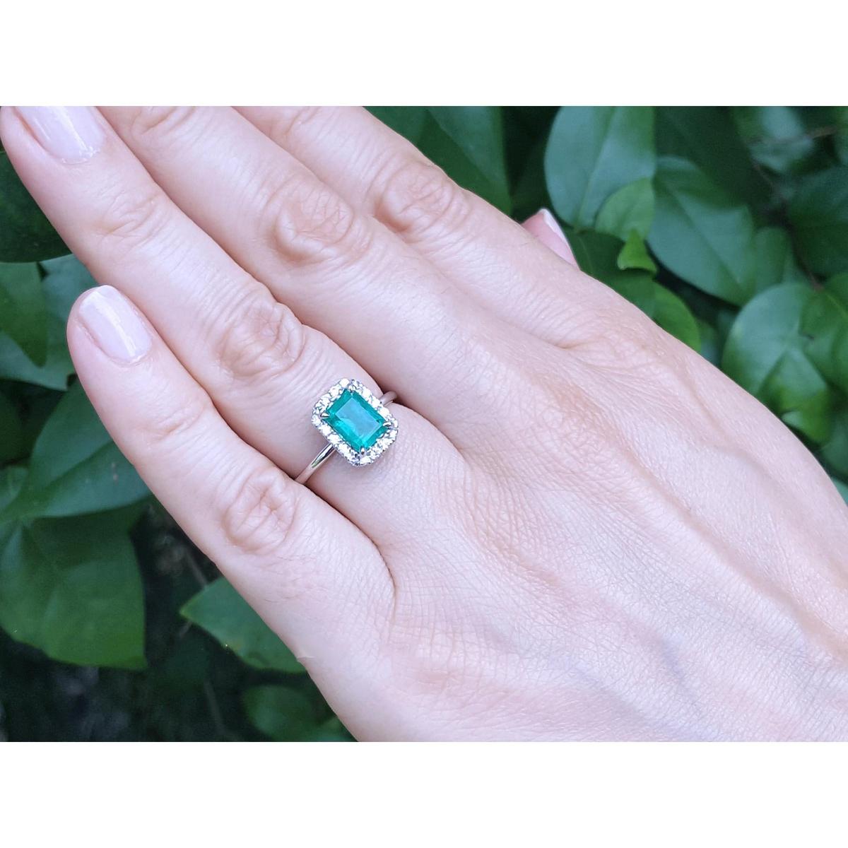 Hand wearing a ring with an emerald stone against a green leafy background. 