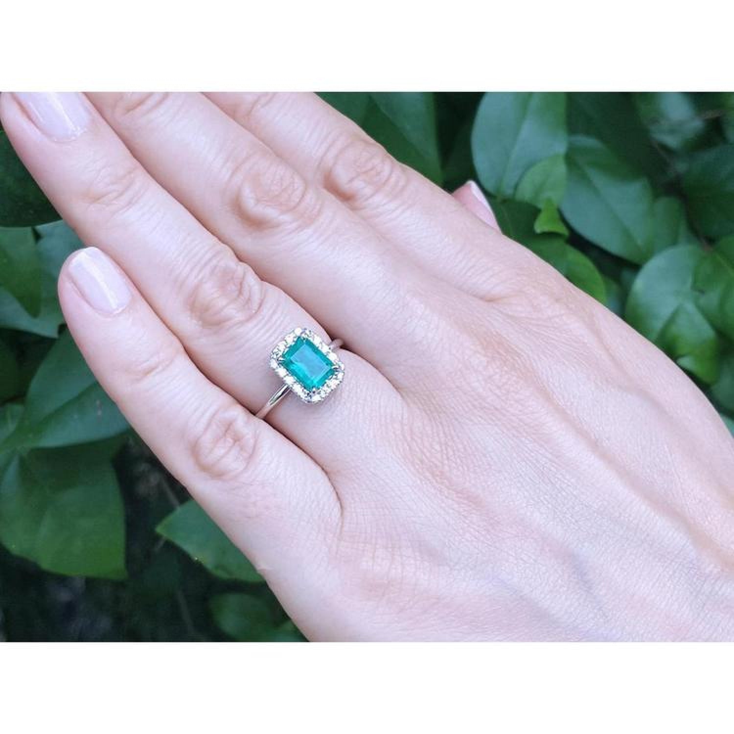 Emerald-cut Muzo emerald ring with halo diamonds in 14K white gold on a hand.