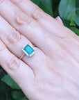 Hand wearing a ring with an emerald stone against a green leafy background. 