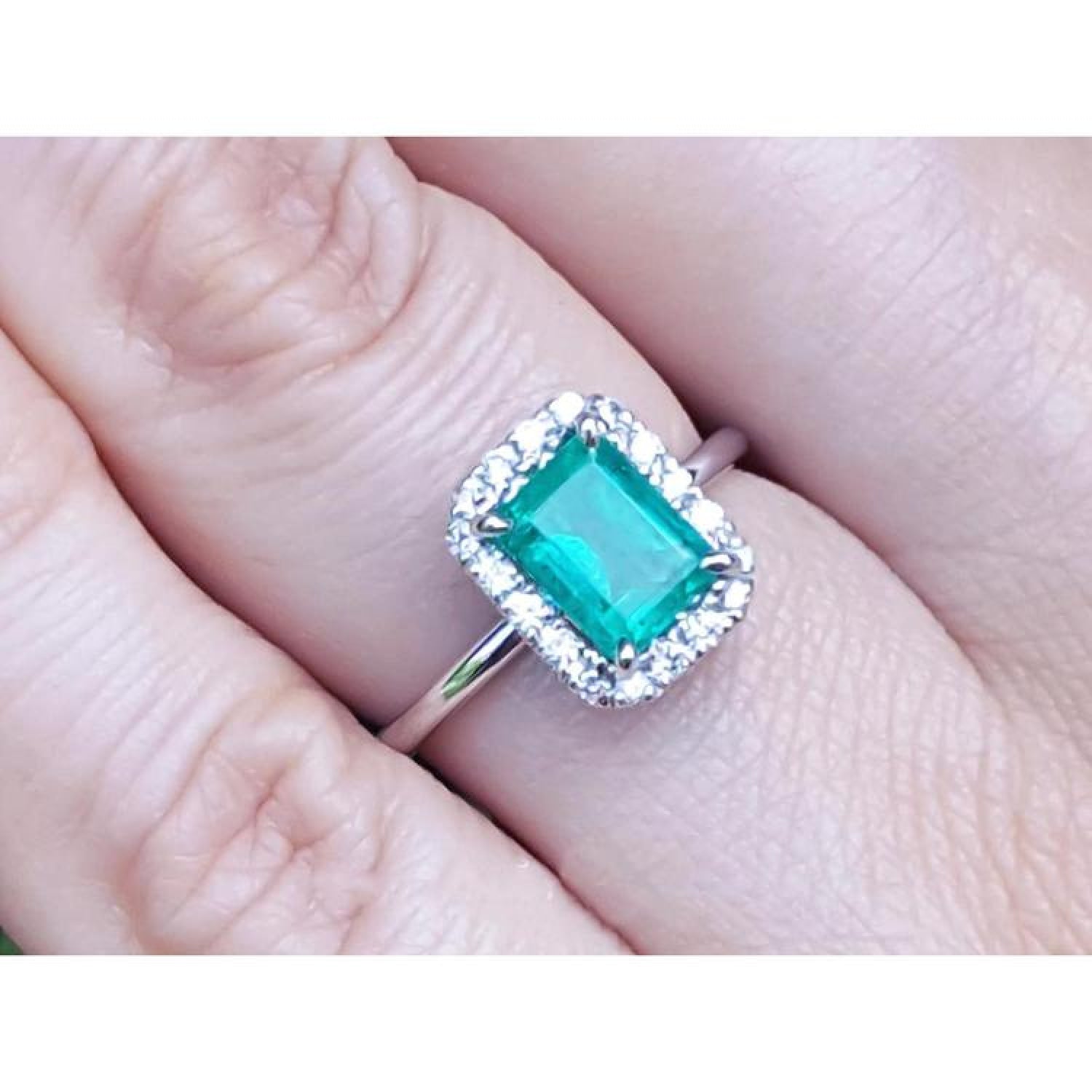 Emerald-cut Muzo emerald halo diamond ring in 14K white gold, worn on a finger.