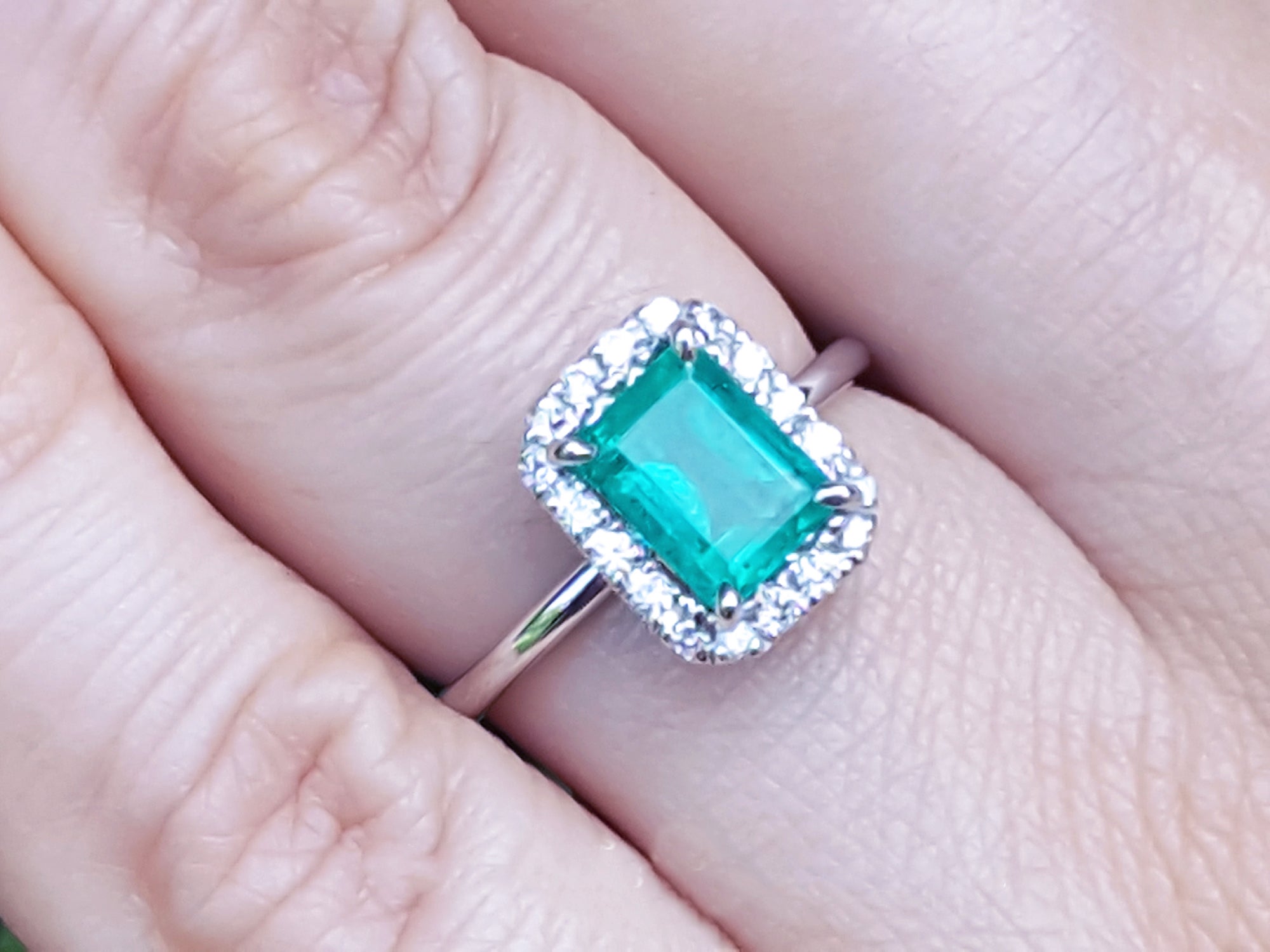Emerald ring with diamond halo on a finger.