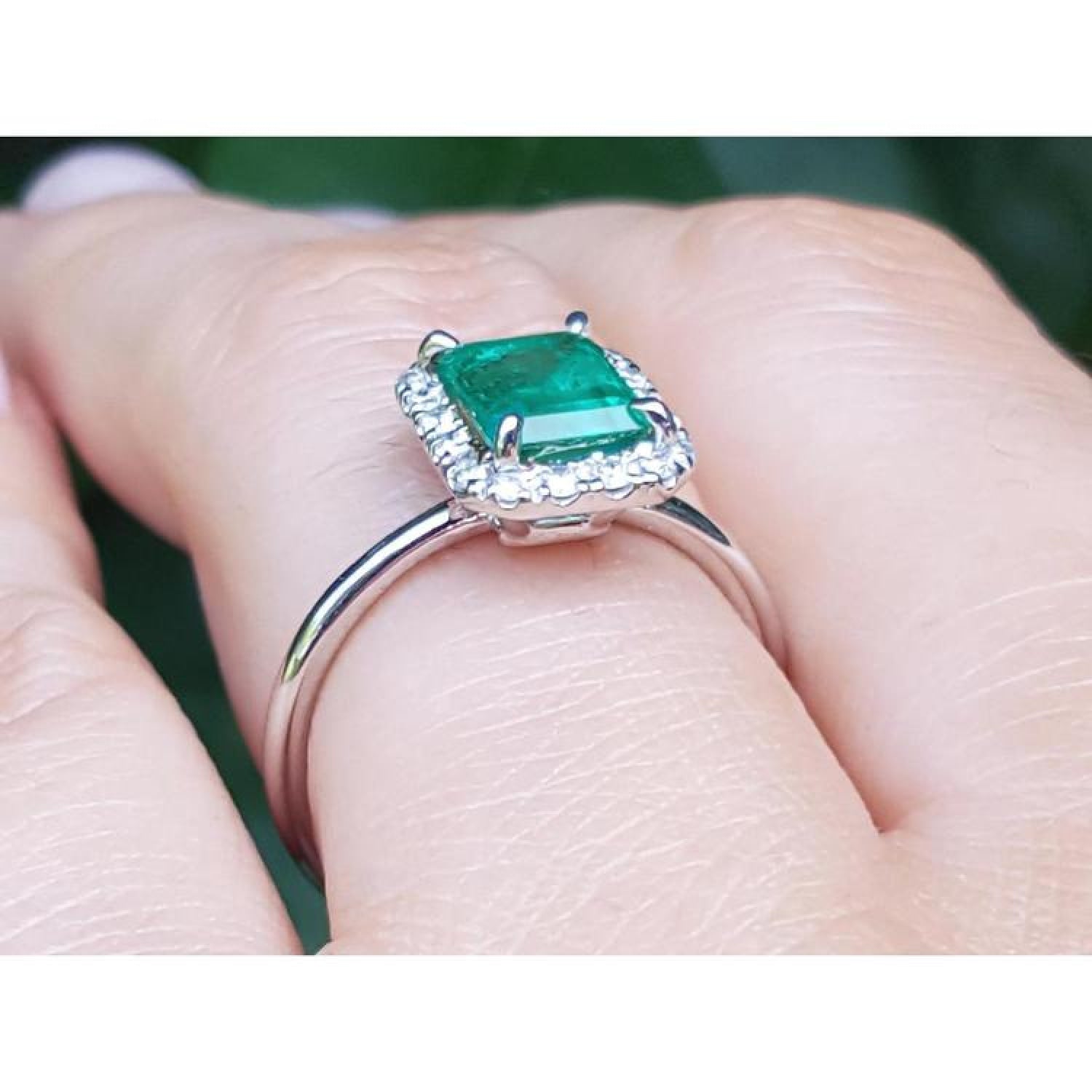 Emerald-cut Muzo emerald halo diamond ring in 14K white gold on a finger.