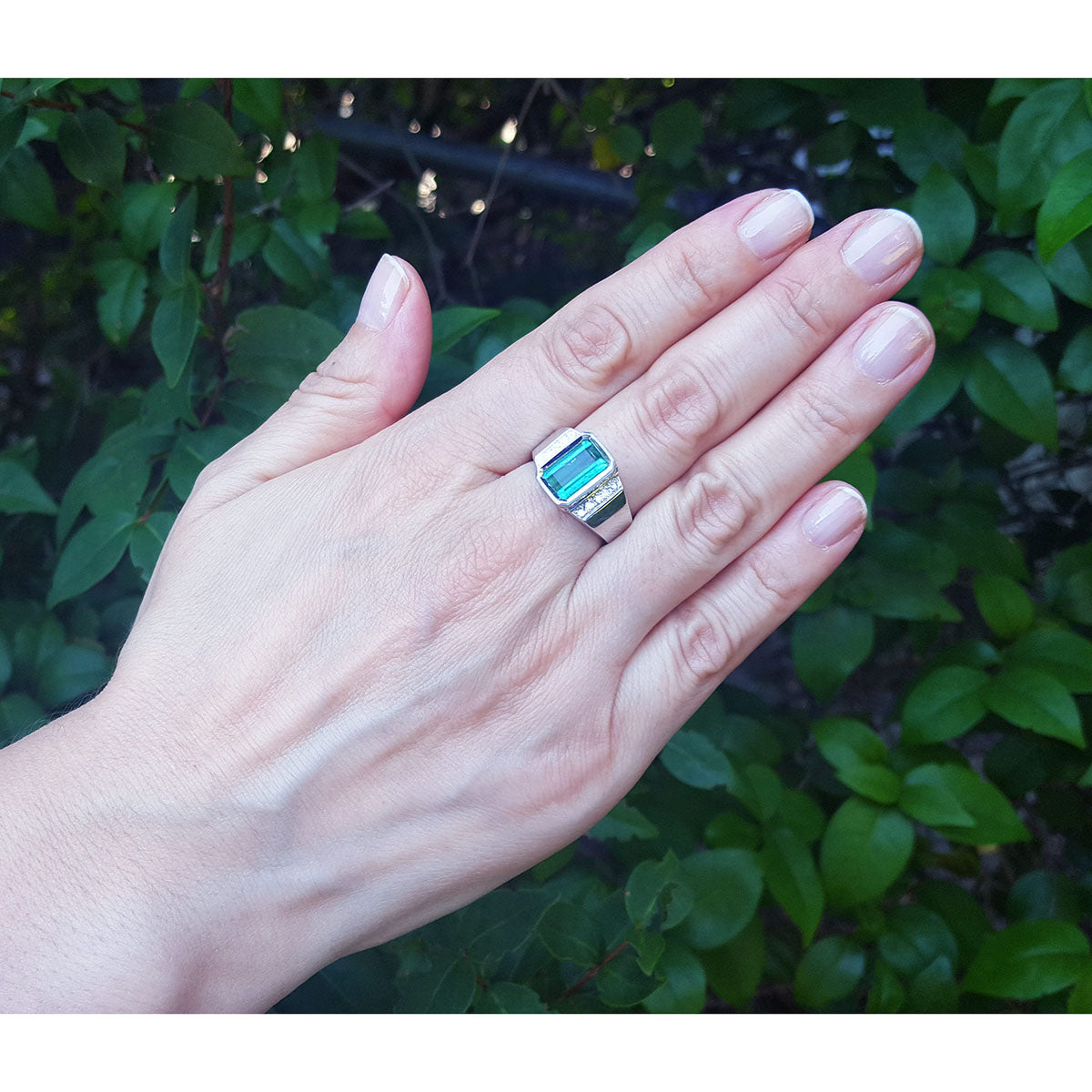 Hand wearing a men&#39;s ring with an emerald-cut emerald against a green leafy background.
