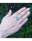 Hand wearing a men's ring with an emerald-cut emerald against a green leafy background.