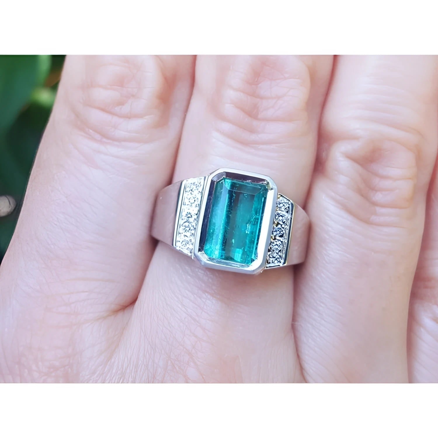 An 18k white gold men’s ring featuring a natural emerald in a bezel setting, accented with diamonds, worn on a finger.