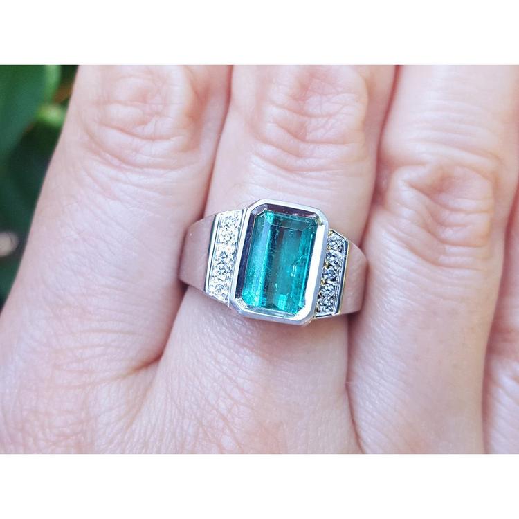An 18k white gold men’s ring featuring a natural emerald in a bezel setting, accented with diamonds, worn on a finger.