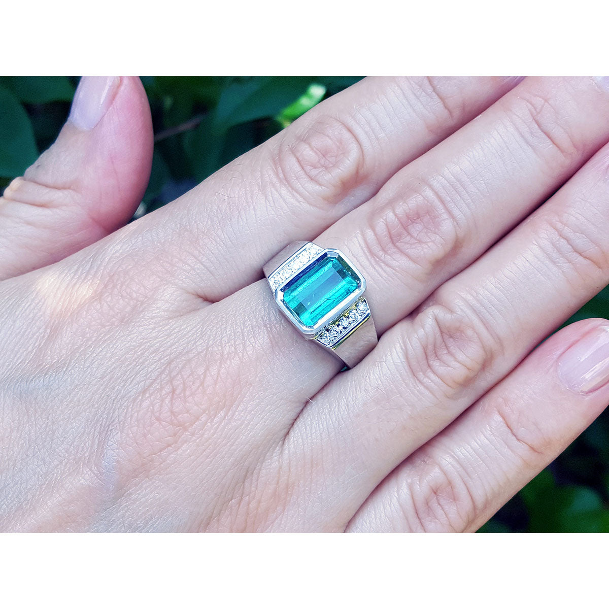 A white gold emerald ring for men on a hand against a blurred green background.