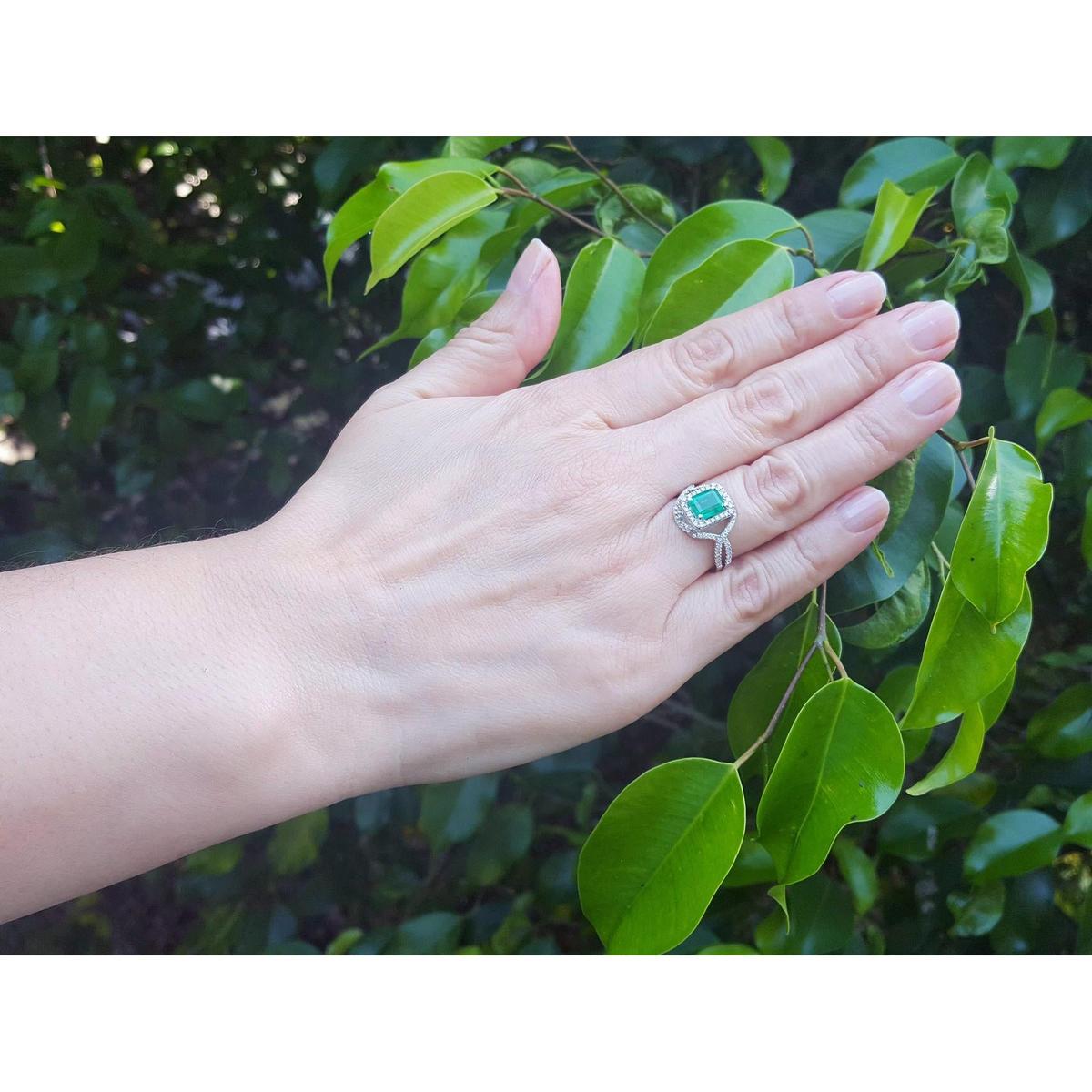 Hand wearing a natural emerald ring with accent diamonds against a leafy background.