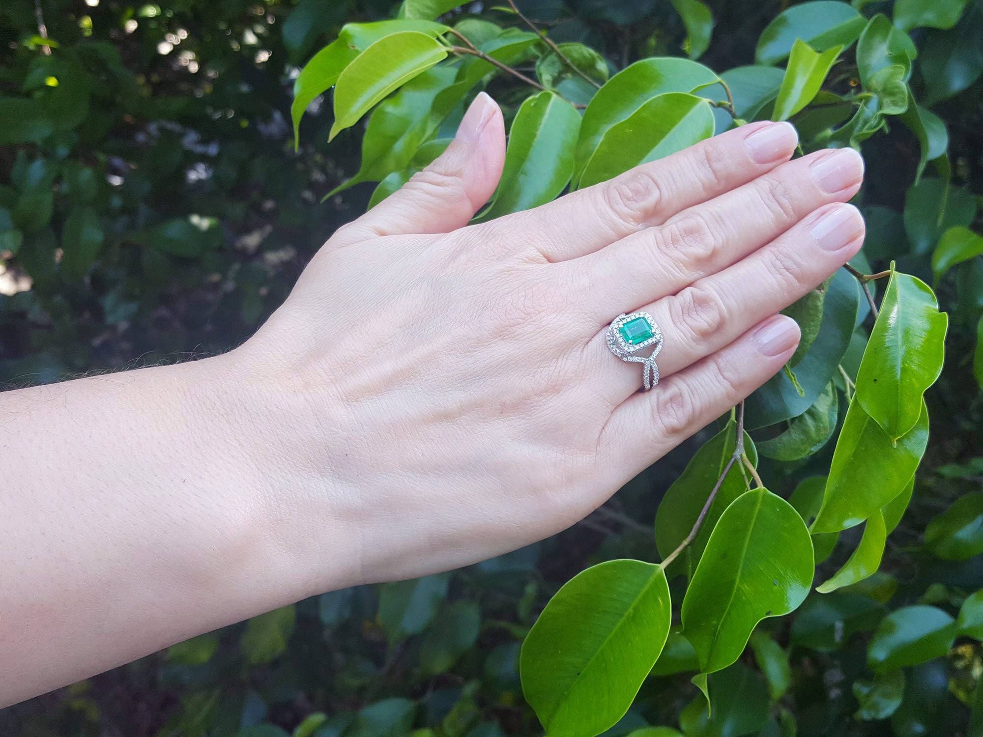 Hand wearing a natural emerald ring with accent diamonds against a leafy background.